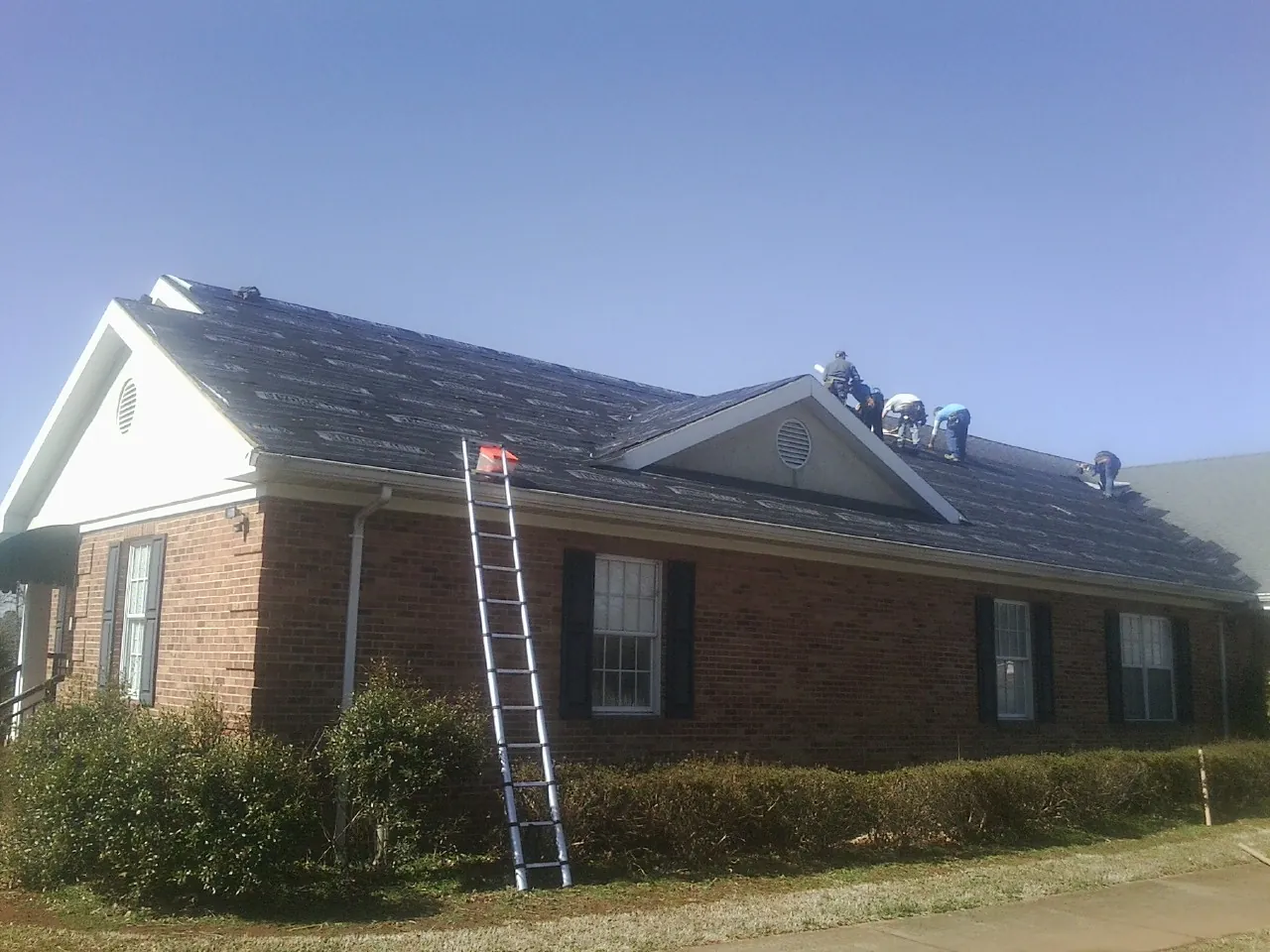 Crew tearing shingles off a brick ranch roof with a ladder set — Metal Roof Repair work by our crew in Branchville